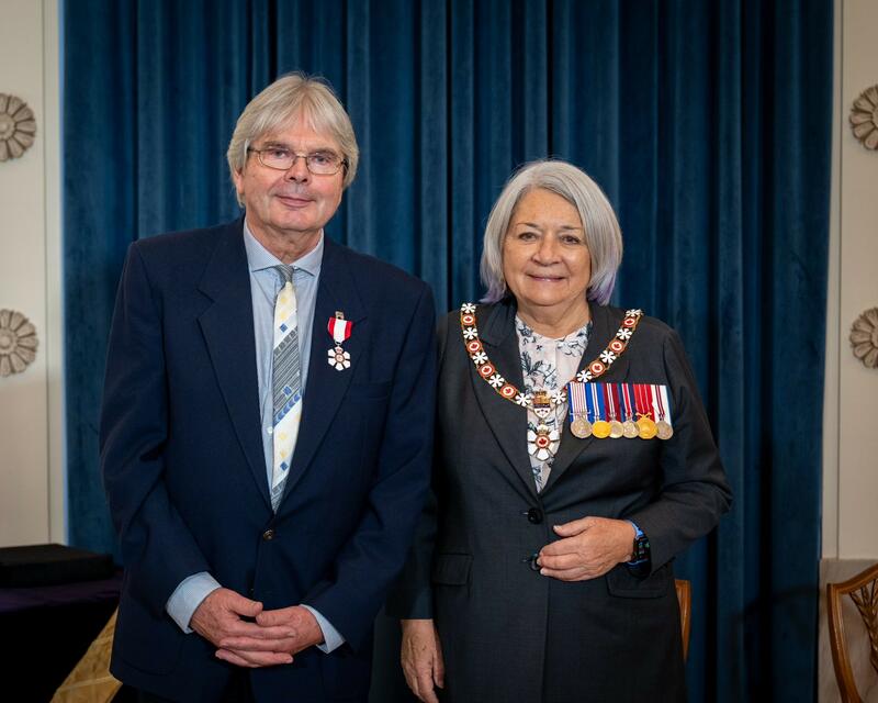 A white-haired man wearing the Order of Canada insignia is smiling to the camera next to Governor General Mary Simon, who is wearing her decorations. Both are standing in front of a blue curtain.