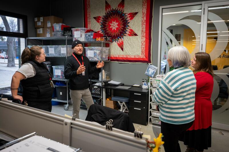 : Governor General Mary Simon is seen from behind, standing and wearing a striped white and green sweater, while she is listening to a gentleman wearing a black beanie hat. Two other women are also listening.