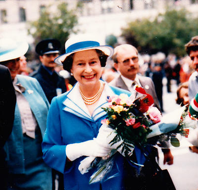 Queen Elizabeth II is walking outside. She is wearing a blue-and-white ensemble and carrying a bouquet of flowers. A crowd looks on.