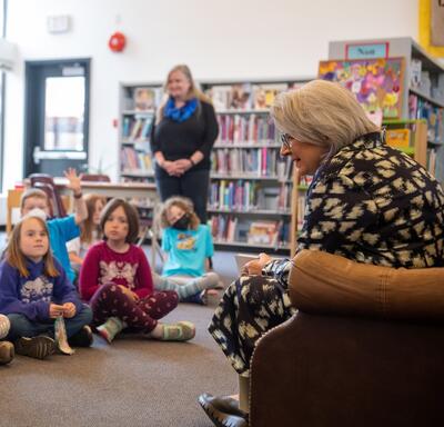 Governor General Simon is reading to a group of children sitting in front of her in a library. She is sitting in a brown armchair. A teacher is standing at the back of the group.