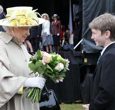 Queen Elizabeth II is wearing a wide-brimmed hat with yellow flowers and a light-tan-coloured trench coat. She holds a bouquet of flowers. A young boy stands in front of her. The Duke of Edinburgh trails behind her. 