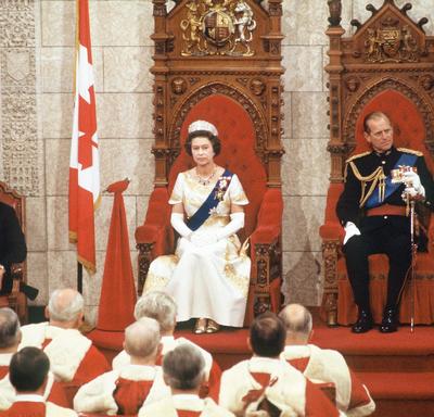 La reine Elizabeth II et le duc d’Édimbourg sont assis chacun sur un trône. La reine porte une robe de couleur crème, une couronne et une écharpe bleu marine. Le duc porte un uniforme militaire complet. Un drapeau du Canada se trouve à leur droite.