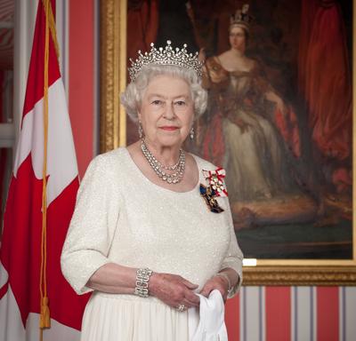 Queen Elizabeth II is dressed in a cream-coloured dress and wears a crown. She is holding a pair of white gloves. She is standing in front of a portrait of Queen Victoria. There is a Canada flag in the background.