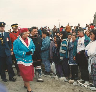 La reine Elizabeth II se déplace devant un groupe d’Inuits. Elle porte une robe de couleur fuchsia, un chapeau assorti, un pardessus bleu et des gants noirs.