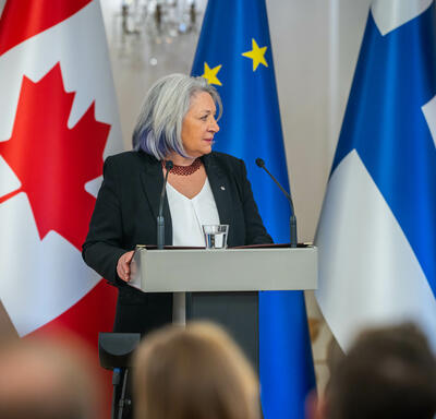 Governor General Simon speaking into a microphone at a podium. Behind her, in order from left to right, are the flags of Canada, Finland and the European Union.  