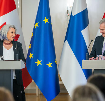 Governor General Simon and President Sauli Niinistö are standing at a podium. Behind them, in order from left to right, are the flags of Canada, Finland and the European Union.  