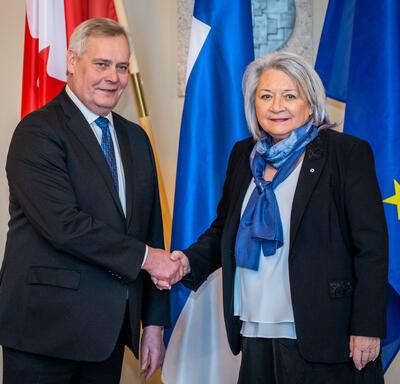Governor General Simon shaking hands with First Deputy Speaker Antti Rinne. Behind them, in order from left to right, are the flags of Canada, Finland and the European Union.  