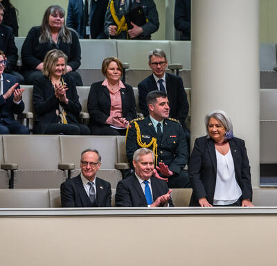 Governor General Simon standing on a balcony in Finland’s Parliament. Several parliamentarians are seated around her. They are clapping.