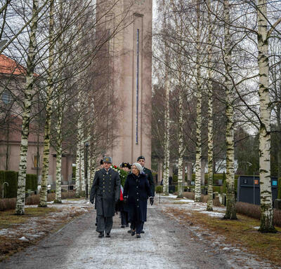 Governor General Simon walking down a gravel path in a cemetery. A man in a uniform is walking beside her and a group of people are walking behind them. Behind the group is a large stone clock tower.