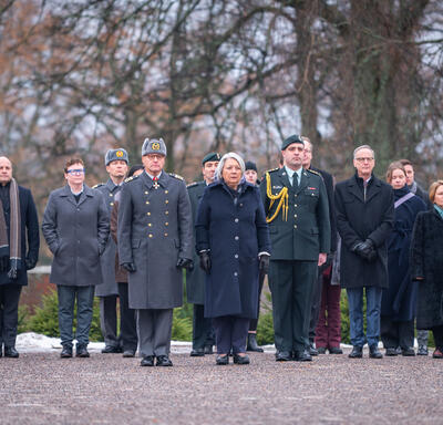 Governor General Simon standing in front of a group of people on a gravel road. Their expressions are sombre. Several people are in military uniforms. 