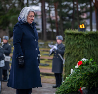 Governor General Simon standing in front of a wreath laid on a black monument. A military band performs behind her. 