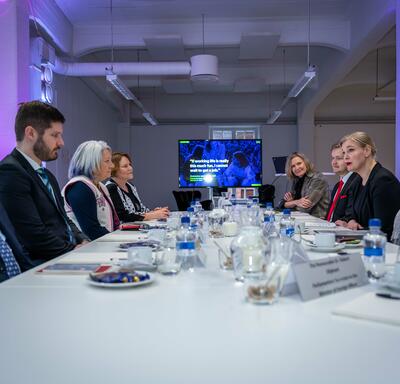 Governor General Simon sitting at a large table with a group of people. A presentation plays on a television at the end of the table.