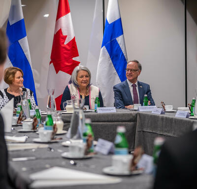 Governor General Simon sitting at the head of a large conference table, in between two other people. Behind her are two flags of Canada and two flags of Finland. Two other people sitting at the table have their backs to the camera.