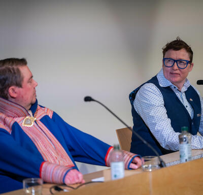 Three people sitting behind a wooden table. They each have a microphone in front of them. The person seated in the middle is speaking. 