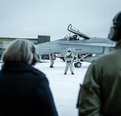 Governor General Simon standing outside next to a man. They are both wearing headsets. One person is standing in the background in front of an airplane.
