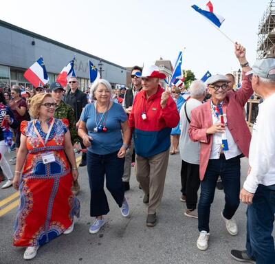 Governor General Mary Simon and Mr. Whit Fraser are walking with a crowd of people outside, participating in National Acadian Day festivities.