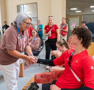 The Governor General of Canada Mary Simon shake hands with a seated Team Canada athlete. Several people, including other athletes and officials, are smiling and observing the interaction.
