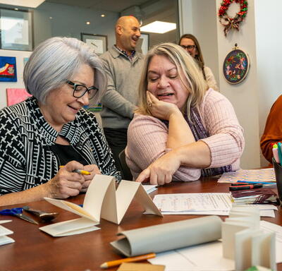 Governor General Mary Simon sitting next to a woman at a table full of craft supplies. The woman is pointing something out to her.