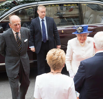 Sa Majesté la Reine et le duc d’Édimbourg sont accueillis par le gouverneur général Johnston et Mme Sharon Johnston. Un garde du corps en costume se tient à côté d’un véhicule en arrière-plan.