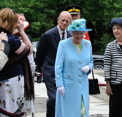 The Queen and the Duke of Edinburgh walk outdoors, along a sidewalk beside a green lawn. The Queen waves. They are accompanied by several men in suits and military uniforms.