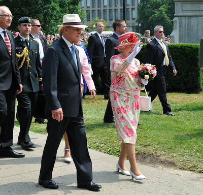 La Reine et le duc d’Édimbourg marchent à l’extérieur, le long d’un trottoir bordé d’une pelouse verte. La Reine fait un signe de la main. Ils sont accompagnés de plusieurs hommes en costume et en uniforme militaire.