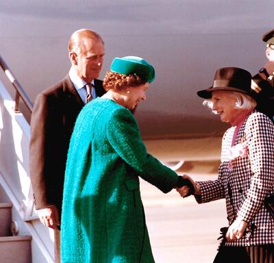 The Queen shakes hands with Governor General Sauvé, who curtsies. The Duke of Edinburgh is standing behind the Queen. A uniformed officer salutes in the background. All are standing at the bottom of a set of stairs extending from an airplane onto the tarm