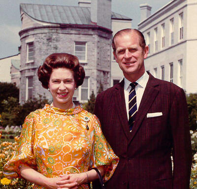 The Queen and the Duke of Edinburgh stand in the private gardens at Rideau Hall. They are both smiling. The Queen is wearing a yellow-and-orange-patterned dress; the Duke is wearing a dark suit.
