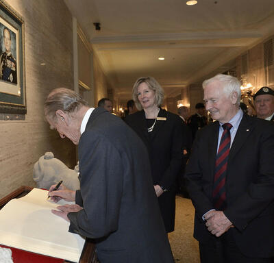 The Duke of Edinburgh writes in a large book as Governor General David Johnston looks on. The pair are in a lobby with other people.