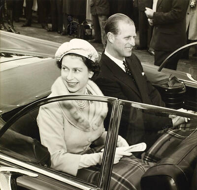 La reine et le duc d’Édimbourg sourient à la foule alors qu’ils sont à bord d’un véhicule décapotable. La photo est en noir et blanc.