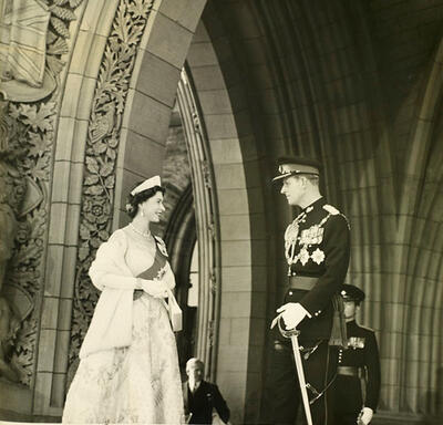 The Queen, dressed in a formal white ball gown and fur wrap, faces the Duke of Edinburgh, who is dressed in a military uniform. The two are standing in a stone archway carved with intricate designs.