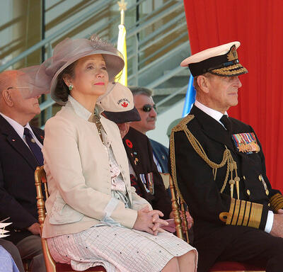 La Reine, la gouverneure générale Clarkson et le duc d’Édimbourg sont assis côte à côte lors d’un événement à l’extérieur. D’autres dignitaires et invités sont assis derrière eux.