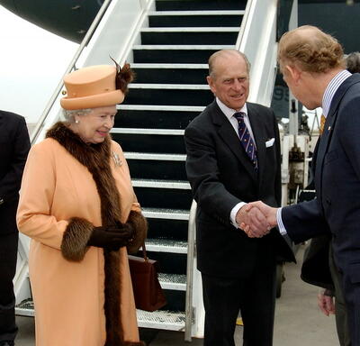 The Duke of Edinburgh shakes hands with John Ralston Saul as The Queen and Governor General Clarkson look on. The group is standing on the tarmac at an airport, at the bottom of a set of stairs leading up to an airplane.