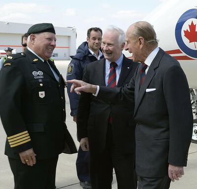 The Duke of Edinburgh laughs as he speaks with Colonel Joe Aitchison, with Governor General David Johnston looking on. The men are standing on the tarmac outside of an airport. A plane bearing the logo of the Canadian Air Force is in the background.