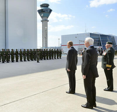 Le duc d’Édimbourg et le gouverneur général Johnston se trouvent sur le tarmac d’un aéroport. Ils font face à un groupe de militaires en uniforme qui se tiennent en formation. À leur droite, le colonel Joe Aitchison dirige le groupe, une épée à la main.
