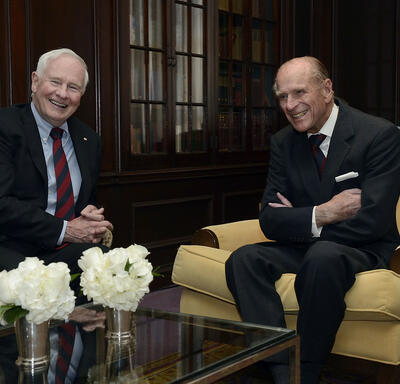 Governor General David Johnston is seated with His Royal Highness the Duke of Edinburgh in a sitting room. Both are smiling. Three vases of white flowers are on the table in the foreground and several shelves of books are in the background.