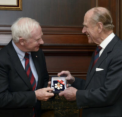 Le gouverneur général David Johnston remet au duc d’Édimbourg une petite boîte contenant l’insigne de l’Ordre du Canada. L’insigne a la forme d’un flocon de neige blanc stylisé avec une feuille d’érable rouge en son centre.