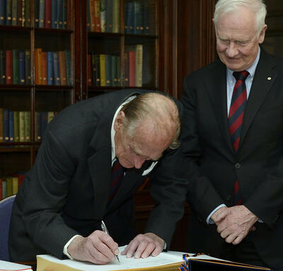 The Duke of Edinburgh signs a large book with gold-leaf edges as Governor General Johnston looks on.