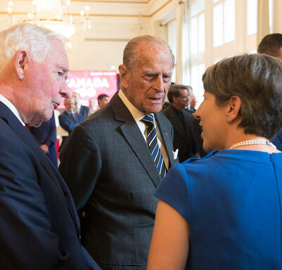 Son Altesse royale le duc d’Édimbourg et le gouverneur général David Johnston parlent à une femme. Ils sont dans une grande salle de réception avec d’autres invités en arrière-plan. Un lustre est suspendu au plafond.