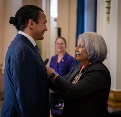 Governor General Simon is smiling to Premier Wab Kinew while pining a Coronation Medal on his vest.