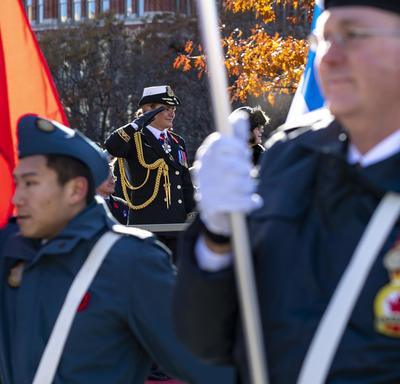Servicemen holding flags are marching in the foreground.  Between their ranks we see the Governor General saluting as well as Ms. Anita Cenerini.