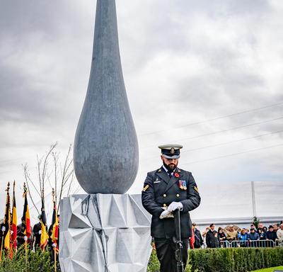 A serviceman stands in front of a monument, eyes towards the ground.  The monument's base has an angular triangle pattern, the top is the shape of a large teardrop. 