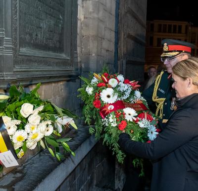 The Governor General of Canada is laying a wreath of flower at a wall monument at night with the help of a military member.
