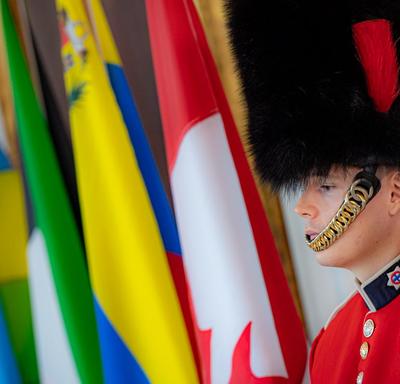 A member of the Governor General’s Foot Guards is standing in front of flags. 