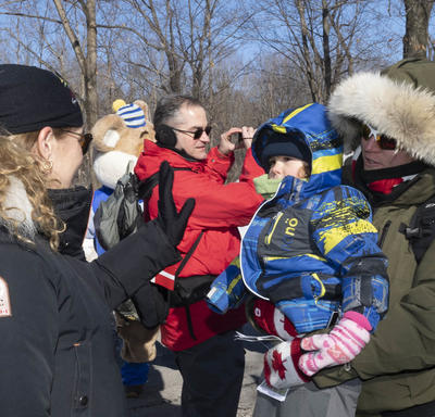 Son Excellence se tient debout devant la caméra et fait signe à un enfant tenu dans les bras de sa mère.  Ils sont debout à l'extérieur en tenue d'hiver. Derrière eux, il y a une foule de gens.