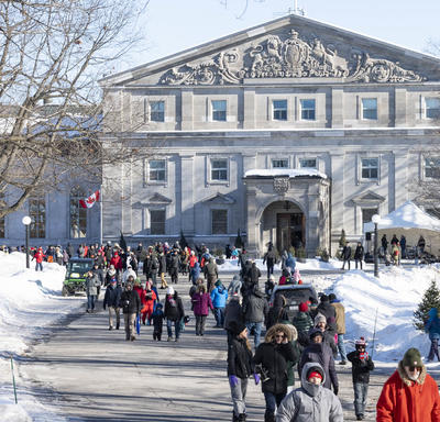 The front façade of Rideau Hall. In front of the residence is a large group of people all dressed in winter gear, walking along the main path.