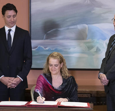 The Governor General signs the oath book. The Prime Minister and the Clerk of the Privy Councillor are on each side of her. 