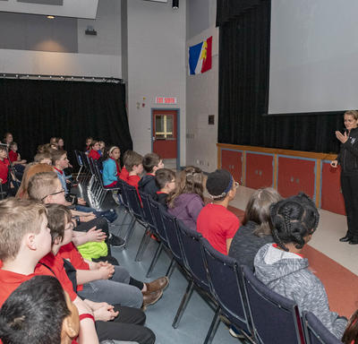 A group of students of grades 1 to 6 are seated in chairs in front of Governor General Julie Payette, during a Power Point presentation.