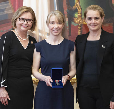A blonde woman is holding a medal in a blue velvet box between Judy May Foote, Lieutenant Governor of Newfoundland and Labrador and Governor General Julie Payette.
