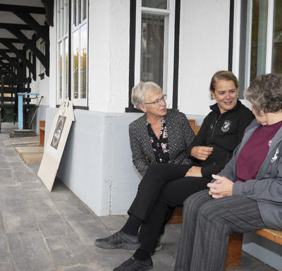 The Governor General is sitting with two women outside of the Cranberry Portage Heritage Museum.