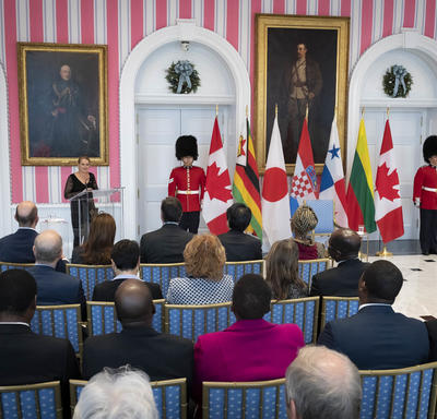 The Governor General delivers remarks at a podium, national flags and a ceremonial guard behind. 